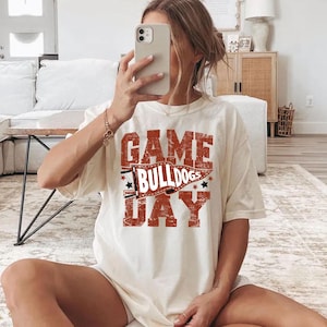 May include: Cream-colored t-shirt with the text "GAME BULLDOGS DAY" in a distressed red font. The design includes a banner and stars. The shirt is a casual, oversized fit.