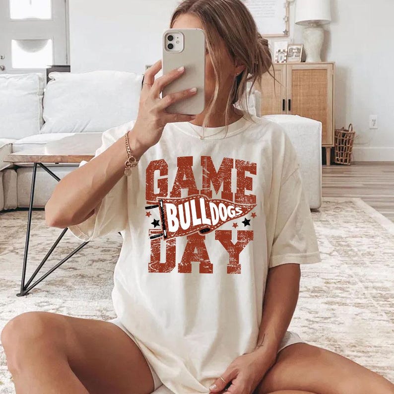 May include: Cream-colored t-shirt with the text "GAME BULLDOGS DAY" in a distressed red font. The design includes a banner and stars. The shirt is a casual, oversized fit.