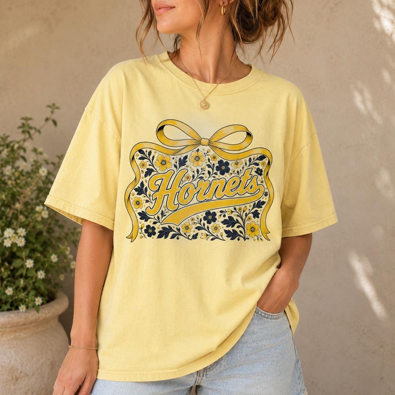May include: A pale yellow short-sleeve t-shirt featuring a graphic print of a large yellow ribbon bow framing the word Hornets in cursive script, surrounded by a detailed pattern of small yellow and dark blue flowers and leafy vines.