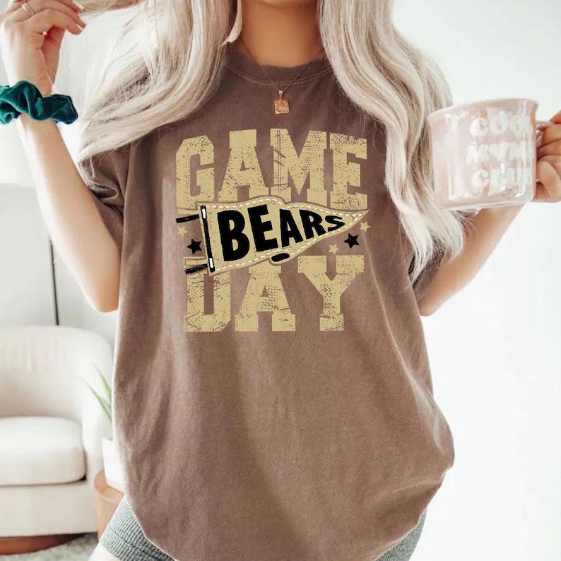 May include: A brown t-shirt with the words "GAME DAY" in a distressed, tan font. The word "BEARS" is inside a pennant. The shirt is a neutral color and has a vintage look. A white mug with text is held in the right hand.
