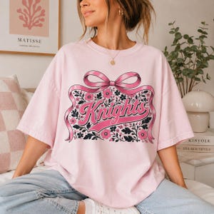 May include: A light pink short-sleeved t-shirt featuring a large graphic of a pink ribbon tied in a bow above the word Knights in a cursive font. The design is surrounded by a pattern of small pink flowers and dark green leaves on a light background.