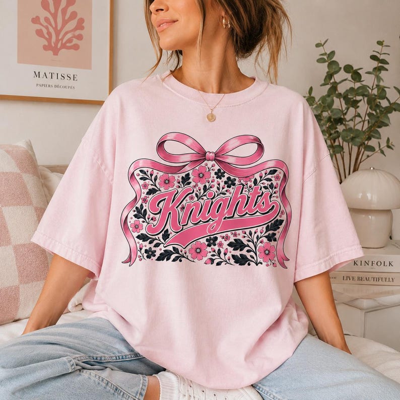 May include: A light pink short-sleeved t-shirt featuring a large graphic of a pink ribbon tied in a bow above the word Knights in a cursive font. The design is surrounded by a pattern of small pink flowers and dark green leaves on a light background.
