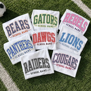 May include: A collection of eight white t-shirts featuring collegiate-style lettering with polka dot patterns. Each shirt displays a unique team name such as Bears, Gators, Rebels, Dawgs, Lions, Panthers, Raiders, or Cougars, followed by the text SCHOOL NAME.