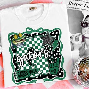 May include: White t-shirt with a green and black graphic. The design includes a cartoon alligator wearing sunglasses, a star, a lightning bolt, and the word "Gators". The shirt also has a "#1" foam finger.