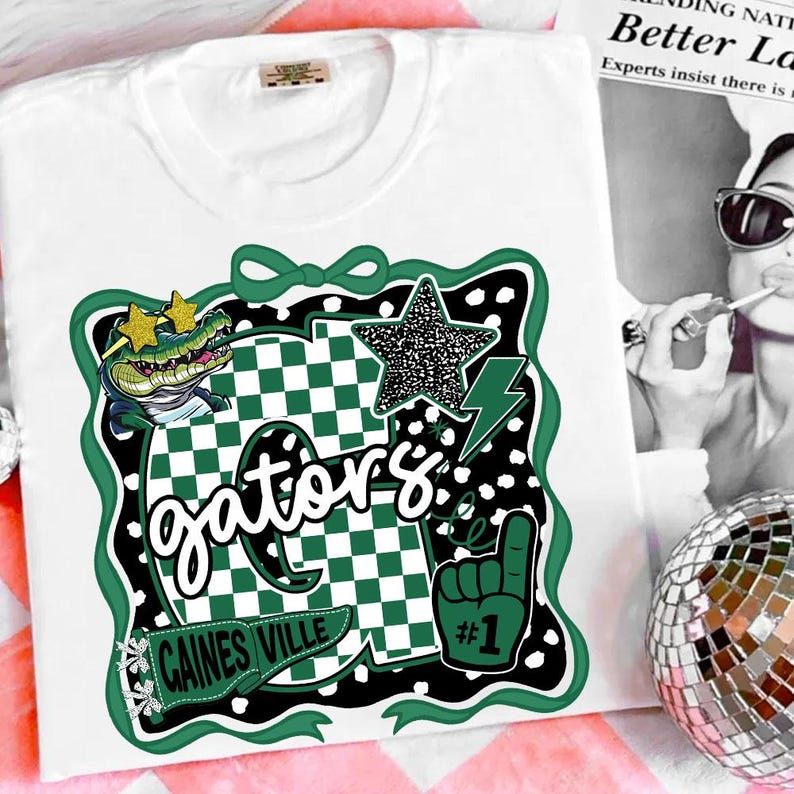 May include: White t-shirt with a green and black graphic. The design includes a cartoon alligator wearing sunglasses, a star, a lightning bolt, and the word "Gators". The shirt also has a "#1" foam finger.