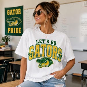 May include: A white short-sleeved t-shirt featuring the green and yellow text LET'S GO GATORS in a collegiate font. The design includes a green and yellow alligator graphic and small green bows on the sides of the word GATORS.
