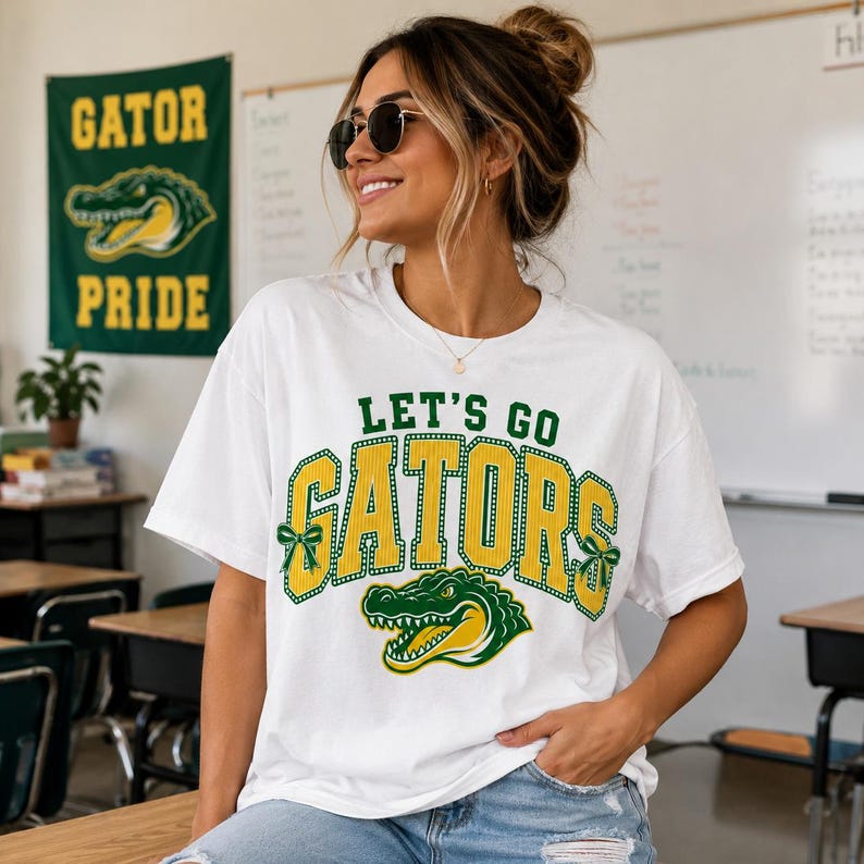 May include: A white short-sleeved t-shirt featuring the green and yellow text LET'S GO GATORS in a collegiate font. The design includes a green and yellow alligator graphic and small green bows on the sides of the word GATORS.
