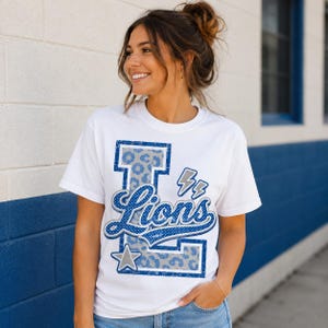 Puede incluir: Una camiseta blanca de manga corta con una letra L grande de color azul con un patr&oacute;n de leopardo gris. La palabra Lions est&aacute; escrita en cursiva azul sobre la letra, acompa&ntilde;ada de dos peque&ntilde;os rayos grises y una estrella gris en la parte inferior.