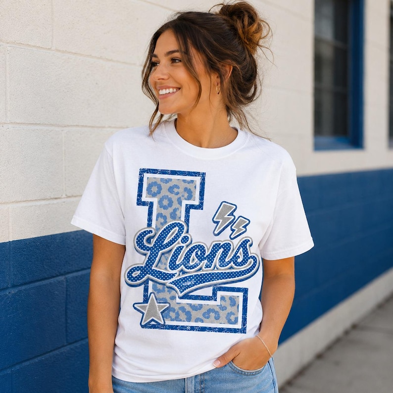 Puede incluir: Una camiseta blanca de manga corta con una letra L grande de color azul con un patr&oacute;n de leopardo gris. La palabra Lions est&aacute; escrita en cursiva azul sobre la letra, acompa&ntilde;ada de dos peque&ntilde;os rayos grises y una estrella gris en la parte inferior.