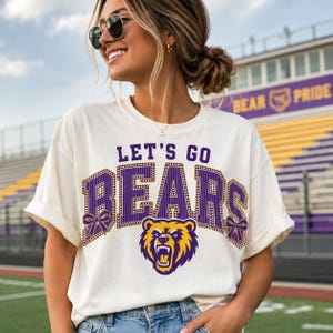 May include: An off-white short-sleeve t-shirt featuring the purple and yellow text LET'S GO BEARS in a collegiate font. The design includes a graphic of a roaring bear head and two purple bows on the sides of the lettering, printed on the chest.
