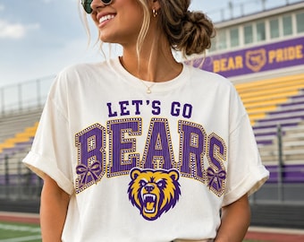 Könnte beinhalten: Ein cremefarbenes Kurzarm-T-Shirt mit dem violetten und gelben Schriftzug LET'S GO BEARS in einer College-Schriftart. Das Design umfasst eine Grafik eines brüllenden Bärenkopfes und zwei violette Schleifen an den Seiten des Schriftzugs auf der Brust.