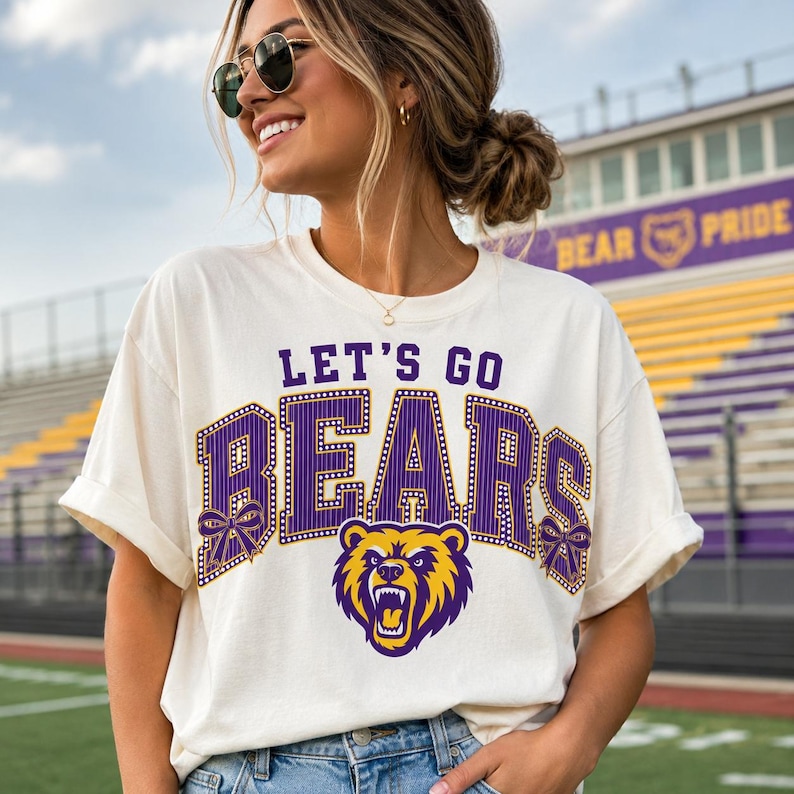 May include: An off-white short-sleeve t-shirt featuring the purple and yellow text LET'S GO BEARS in a collegiate font. The design includes a graphic of a roaring bear head and two purple bows on the sides of the lettering, printed on the chest.