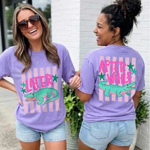 Peut inclure: Deux personnes portant des t-shirts lavande avec un motif d'alligator de dessin animé. Le devant du t-shirt dit "LATER" avec un alligator vert et des rayures roses. Le dos du t-shirt dit "AFTER WHILE" avec un alligator vert et des rayures roses.