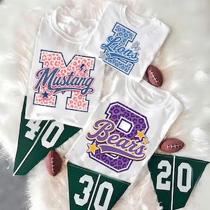 Puede incluir: Tres camisetas blancas con letras grandes en bloque con patrones de estampado animal y nombres de equipos. Una camiseta muestra una M con estampado de leopardo rosa con Mustang, otra una L azul con Lions, y la tercera una B morada con Bears.