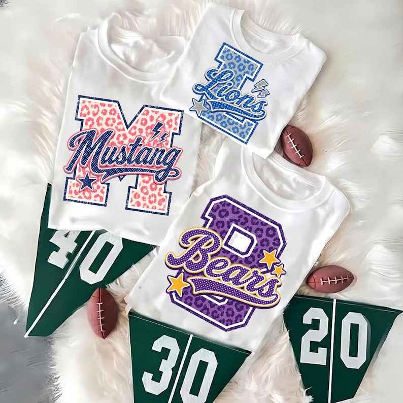 Puede incluir: Tres camisetas blancas con letras grandes en bloque con patrones de estampado animal y nombres de equipos. Una camiseta muestra una M con estampado de leopardo rosa con Mustang, otra una L azul con Lions, y la tercera una B morada con Bears.