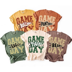 May include: Five short-sleeved t-shirts in various colors, each with a distressed graphic. The shirts feature the words "Game Day" above a team name, such as "Bulldogs," "Panthers," "Eagles," "Tigers," and "Bears." The text is in a collegiate style.