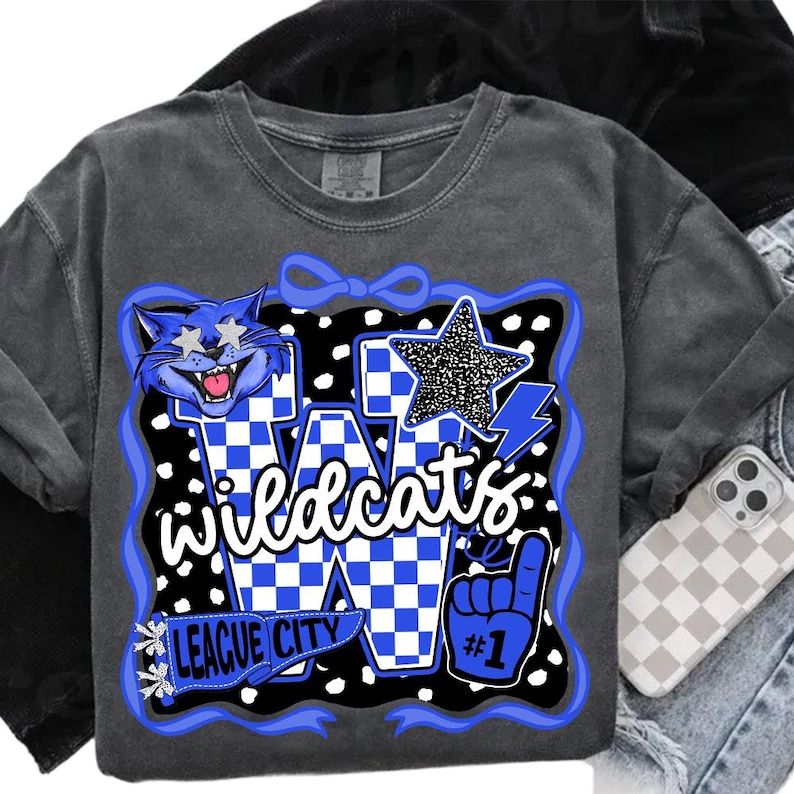 May include: A grey Wildcats sweatshirt featuring a blue and white graphic design. The graphic includes a cartoon cat, a star, and the words "Wildcats" and "League City". The design is outlined in blue and has a black and white polka dot background.