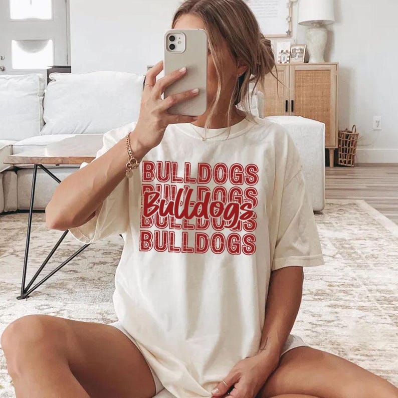 Pu&ograve; includere: T-shirt color crema con la scritta "BULLDOGS" in rosso, ripetuta in un design impilato. La maglietta ha maniche corte e una vestibilit&agrave; ampia. La persona tiene in mano un telefono.