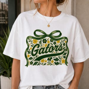 May include: A white short-sleeve t-shirt featuring a graphic print of a large green ribbon bow tied around a cluster of yellow and green flowers and leaves. The word Gators is written in a dark green cursive script across the center of the design.