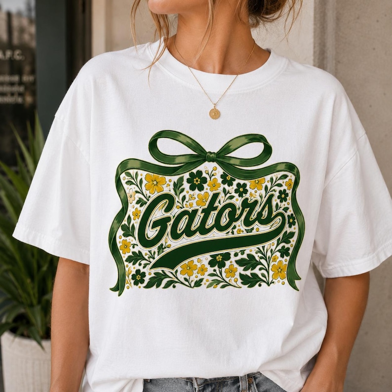 May include: A white short-sleeve t-shirt featuring a graphic print of a large green ribbon bow tied around a cluster of yellow and green flowers and leaves. The word Gators is written in a dark green cursive script across the center of the design.