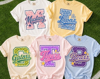 Könnte beinhalten: Fünf kurzärmelige T-Shirts mit Rundhalsausschnitt in Weiß, Gelb, Creme, Hellrosa und Hellblau. Jedes Shirt zeigt einen großen College-Buchstaben mit Leopardenmuster und einen Teamnamen in Schreibschrift: Mustang, Lions, Gators, Tigers und Bears.