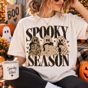 May include: A cream-coloured t-shirt featuring the text SPOOKY SEASON in black distressed lettering. The design includes three ghosts with different patterns, each holding a black cat, accented by small orange stars and black hearts.