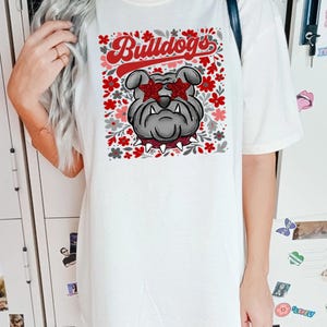 以下が含まれることがあります： 白いTシャツ。星型のサングラスをかけたブルドッグのグラフィックと、赤いスクリプトで「Bulldogs」の文字が描かれています。デザインには、赤、ピンク、グレーの花柄が含まれています。このシャツはカジュアルなユニセックススタイルです。