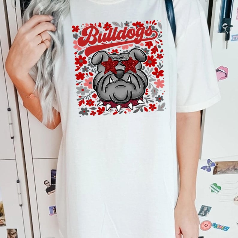 以下が含まれることがあります： 白いTシャツ。星型のサングラスをかけたブルドッグのグラフィックと、赤いスクリプトで「Bulldogs」の文字が描かれています。デザインには、赤、ピンク、グレーの花柄が含まれています。このシャツはカジュアルなユニセックススタイルです。