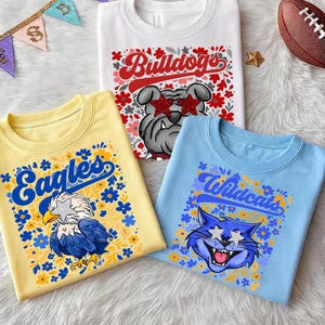 以下が含まれることがあります： チームのマスコットデザインのTシャツ3枚。白いシャツには、星型のメガネをかけたブルドッグと「Bulldogs」の文字が描かれています。黄色のシャツにはワシと「Eagles」の文字が描かれています。青いシャツには猫と「Wildcats」の文字が描かれています。