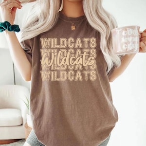 Pu&ograve; includere: Una maglietta marrone con la parola "WILDCATS" stampata in un design impilato in un colore marrone pi&ugrave; chiaro. La maglietta ha maniche corte e girocollo. Una tazza con testo &egrave; tenuta nella mano destra.