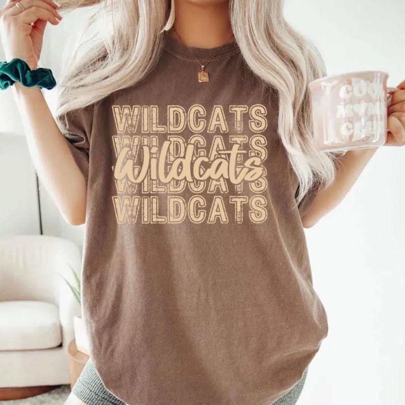Pu&ograve; includere: Una maglietta marrone con la parola "WILDCATS" stampata in un design impilato in un colore marrone pi&ugrave; chiaro. La maglietta ha maniche corte e girocollo. Una tazza con testo &egrave; tenuta nella mano destra.