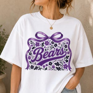 May include: A white short-sleeve t-shirt featuring a graphic print of a large purple ribbon tied in a bow above the word Bears in purple script. The design includes purple and dark blue floral illustrations surrounding the text.