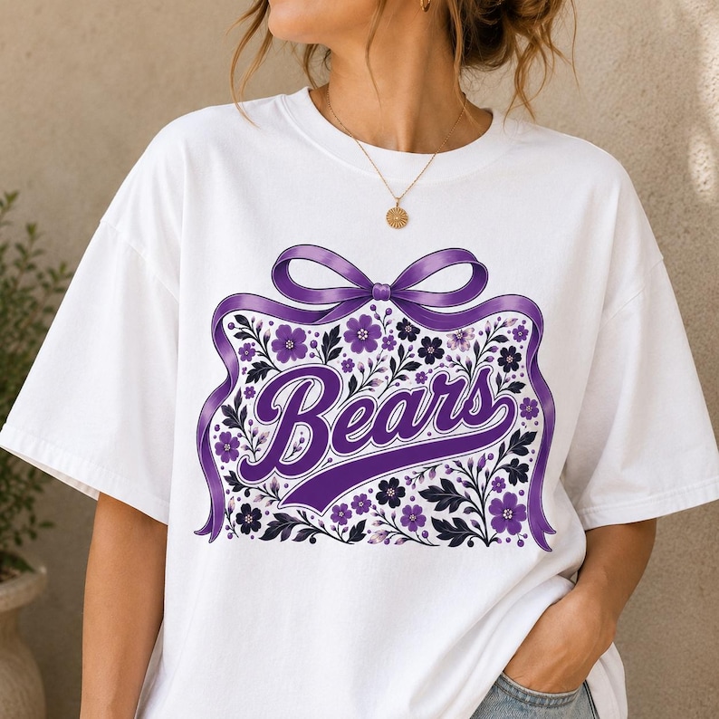 May include: A white short-sleeve t-shirt featuring a graphic print of a large purple ribbon tied in a bow above the word Bears in purple script. The design includes purple and dark blue floral illustrations surrounding the text.