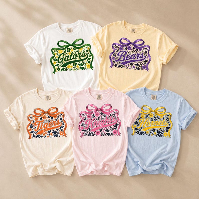 May include: Five short-sleeve t-shirts in white, yellow, cream, light pink, and light blue. Each shirt features a central graphic of a ribbon bow above a floral rectangular frame containing the text Gators, Bears, Tigers, Knights, or Hornets.