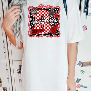 May include: White t-shirt with a graphic design featuring the word "Bulldogs". The design includes a bulldog, a star, a foam finger, and the words "Karns City". The design is in red, black, and white.