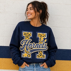 Puede incluir: Una sudadera azul marino de cuello redondo con una letra H grande con un patr&oacute;n de leopardo amarillo y negro. La palabra Hornets est&aacute; impresa en letra cursiva sobre la letra, acompa&ntilde;ada por dos rayos amarillos y una estrella amarilla.