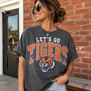May include: A charcoal gray short-sleeve t-shirt featuring the text LET'S GO TIGERS in white and orange block letters. The design includes a graphic of a tiger face and two dark blue bows on the sides of the text, all printed on the front.