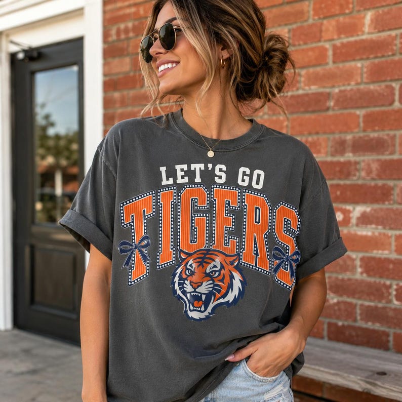 May include: A charcoal gray short-sleeve t-shirt featuring the text LET'S GO TIGERS in white and orange block letters. The design includes a graphic of a tiger face and two dark blue bows on the sides of the text, all printed on the front.