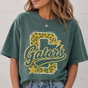 Puede incluir: Una camiseta de manga corta de color verde oscuro con un gran estampado gr&aacute;fico de una letra G en bloque con un patr&oacute;n de leopardo amarillo y verde. La palabra Gators est&aacute; escrita en una fuente cursiva blanca con un borde verde, acentuada por una estrella amarilla.