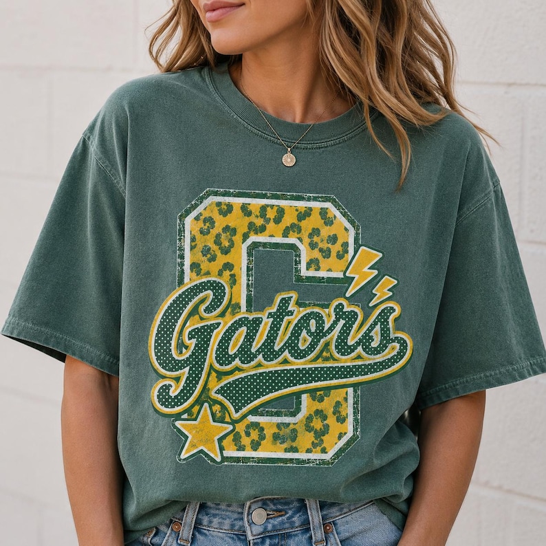 Puede incluir: Una camiseta de manga corta de color verde oscuro con un gran estampado gr&aacute;fico de una letra G en bloque con un patr&oacute;n de leopardo amarillo y verde. La palabra Gators est&aacute; escrita en una fuente cursiva blanca con un borde verde, acentuada por una estrella amarilla.