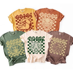 May include: Five short-sleeved t-shirts in various colours, each featuring a team mascot and name. The shirts include a yellow "Bulldogs" shirt, a rust-coloured "Panthers" shirt, a green "Bears" shirt, a cream "Tigers" shirt, and a brown "Eagles" shirt.