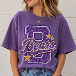 Puede incluir: Una camiseta de manga corta morada con una letra B grande con estampado de leopardo morado. La palabra Bears est&aacute; escrita en letra cursiva con puntos blancos y un borde dorado. Dos estrellas doradas est&aacute;n situadas cerca de la letra B.