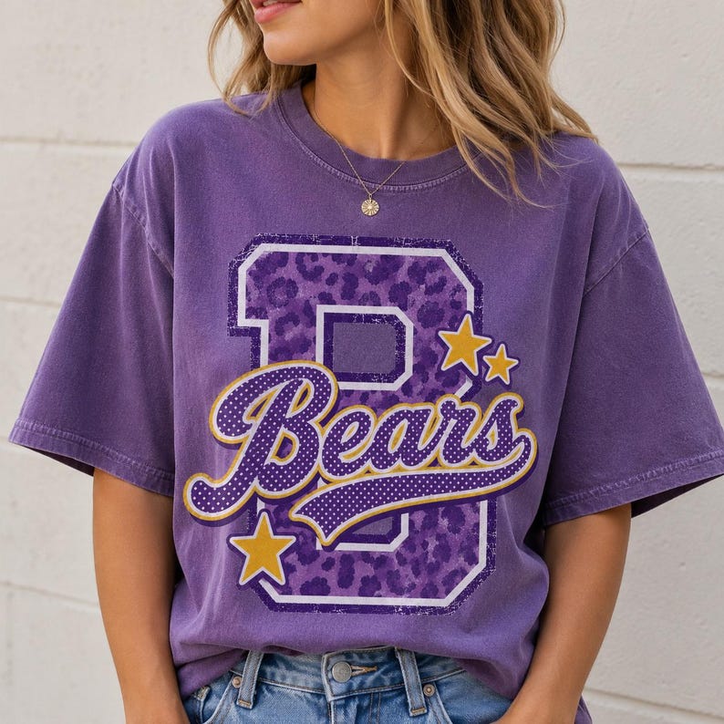 Puede incluir: Una camiseta de manga corta morada con una letra B grande con estampado de leopardo morado. La palabra Bears est&aacute; escrita en letra cursiva con puntos blancos y un borde dorado. Dos estrellas doradas est&aacute;n situadas cerca de la letra B.