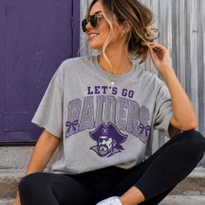 May include: A heather gray short-sleeve t-shirt featuring the text LET'S GO RAIDERS in purple and white block letters with a dotted outline. A graphic of a pirate wearing a purple hat with an R emblem is centered below the text.