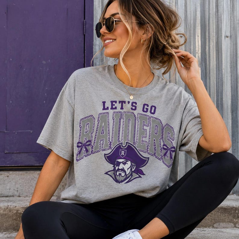 May include: A heather gray short-sleeve t-shirt featuring the text LET'S GO RAIDERS in purple and white block letters with a dotted outline. A graphic of a pirate wearing a purple hat with an R emblem is centered below the text.