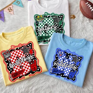 May include: Three t-shirts in white, yellow, and blue, each with a unique graphic design. The white shirt features a green and black design with the word "gators". The yellow shirt has a red and black design with the word "bulldogs". The blue shirt has a blue and black design with the word "wildcats".