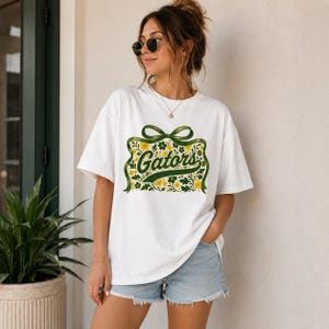 May include: A white short-sleeve t-shirt featuring a central graphic of a green ribbon tied in a bow above a rectangular floral pattern in shades of green and yellow, with the word Gators written in a cursive green font across the center.