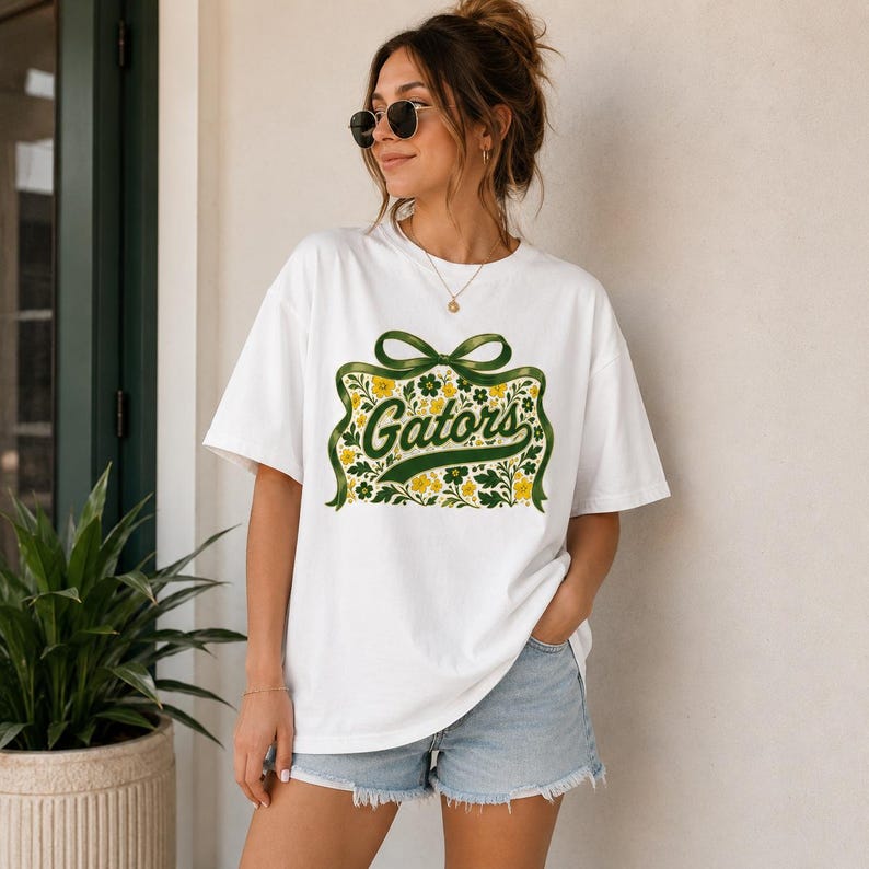 May include: A white short-sleeve t-shirt featuring a central graphic of a green ribbon tied in a bow above a rectangular floral pattern in shades of green and yellow, with the word Gators written in a cursive green font across the center.