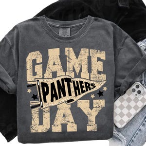 May include: Dark gray sweatshirt with the words "GAME DAY PANTHERS" in a distressed, tan font. The word "PANTHERS" is inside a banner. Black stars are on either side of the banner. The sweatshirt is folded with a phone case visible.