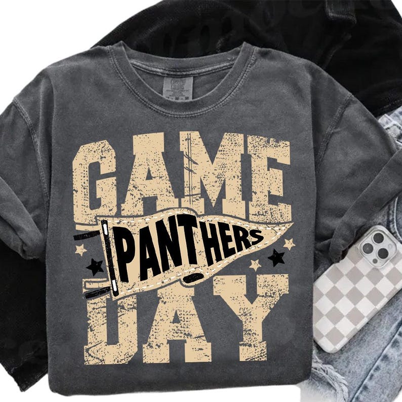 May include: Dark gray sweatshirt with the words "GAME DAY PANTHERS" in a distressed, tan font. The word "PANTHERS" is inside a banner. Black stars are on either side of the banner. The sweatshirt is folded with a phone case visible.